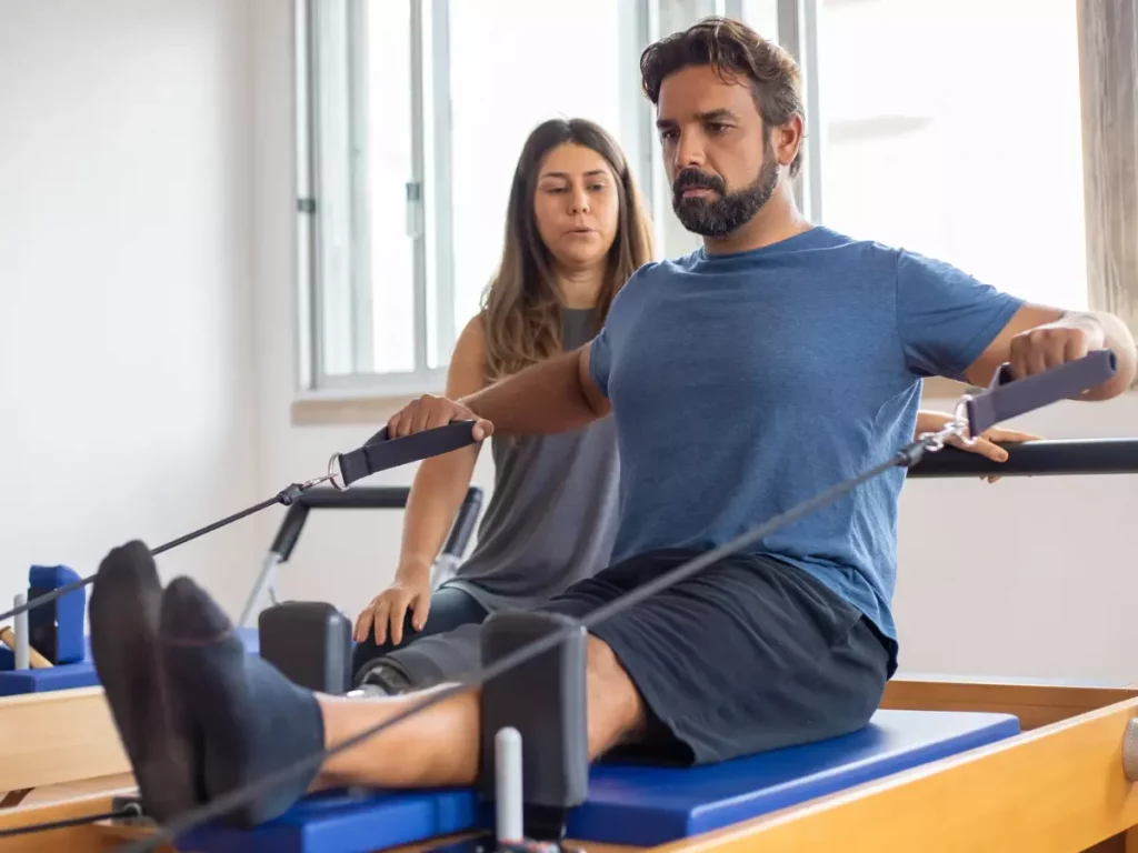 Homem praticando pilates, sentado no Reformer, e ao seu lado, está sua instrutora acompanhando sua prática.